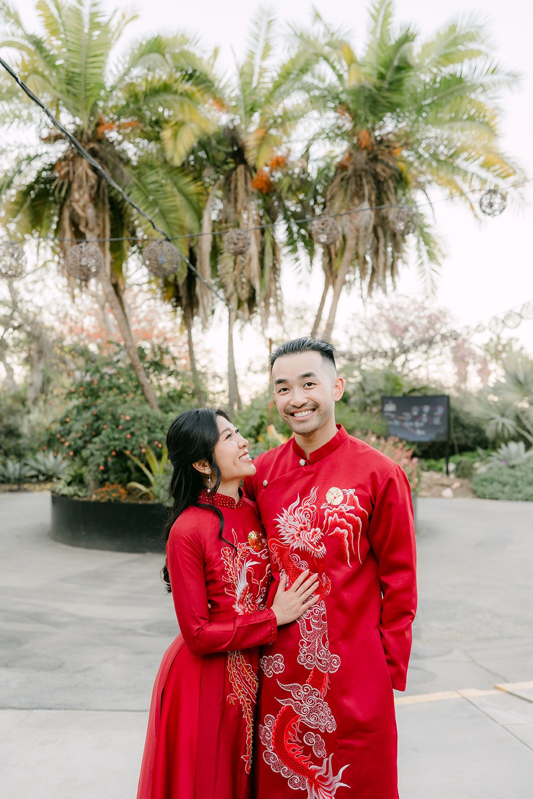 Kim leaning on Vincent's shoulder in their red ao dai among the palms.