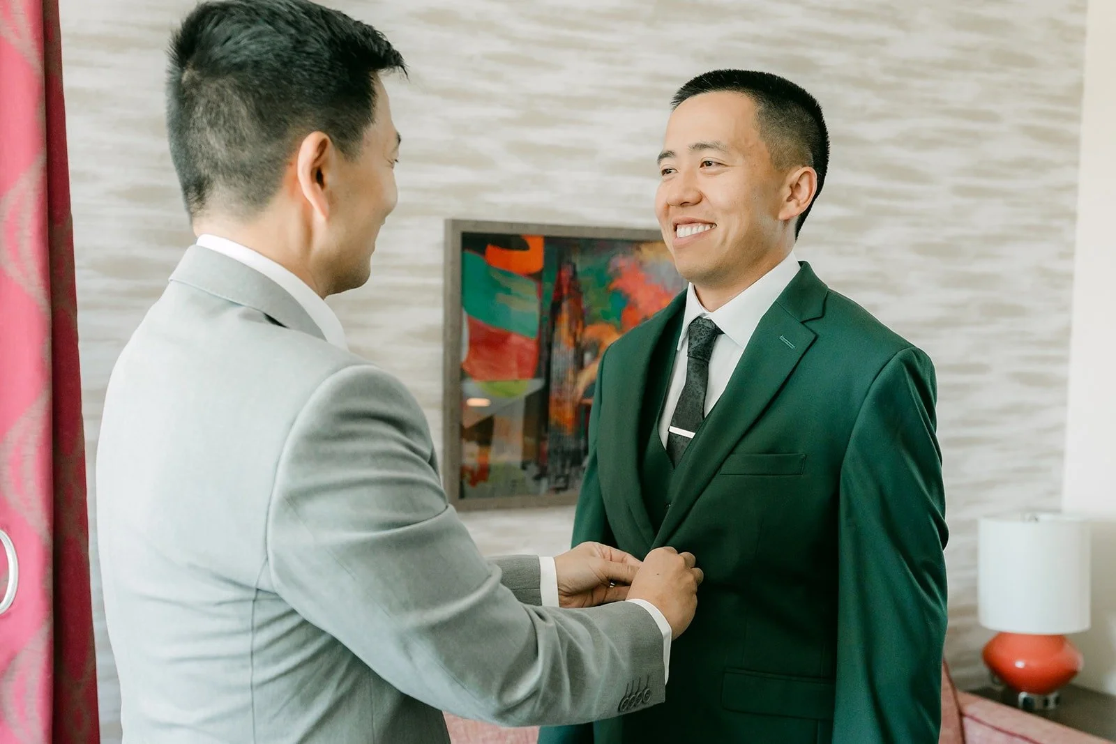 A groomsman fixes Kevin's dark green suit jacket at Home2 Suites