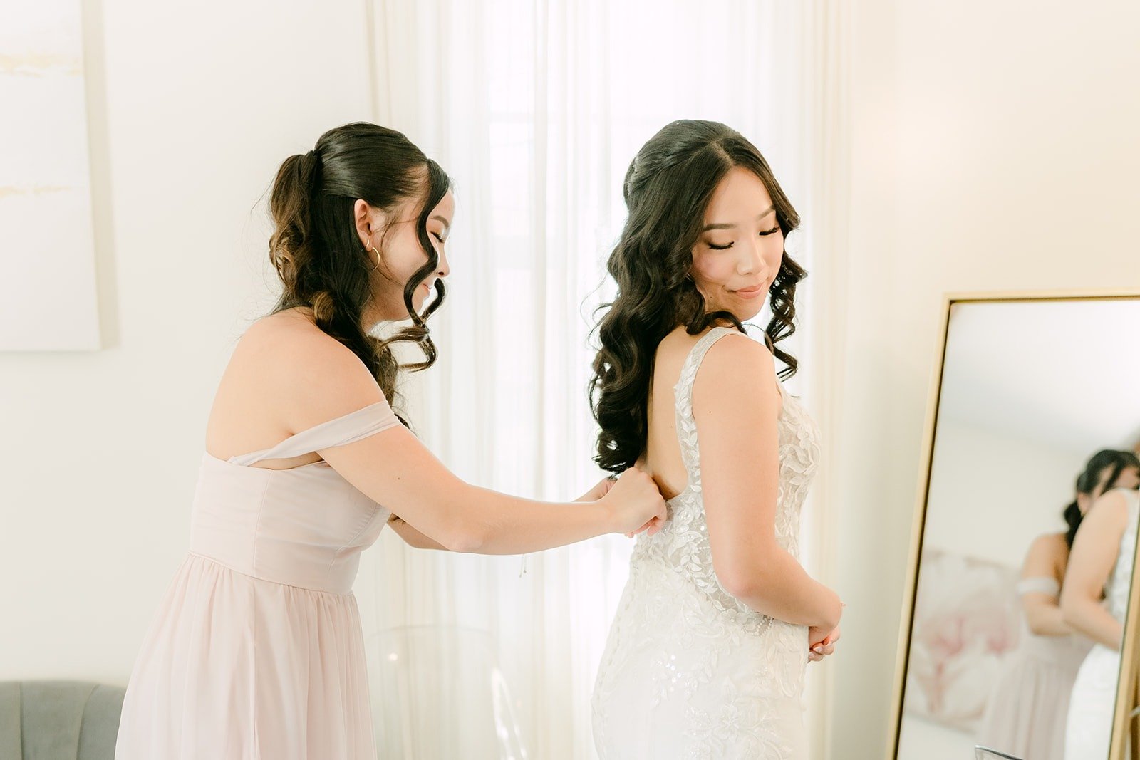 A bridesmaid in blush fastening the back of Kelli's lace gown by the window