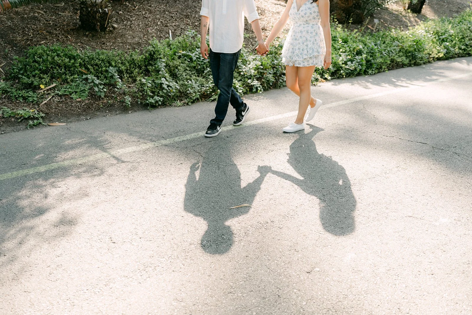 Francesca and Daniel walk hand in hand down a sunlit garden path at the LA Arboretum, their shadows stretching ahead.