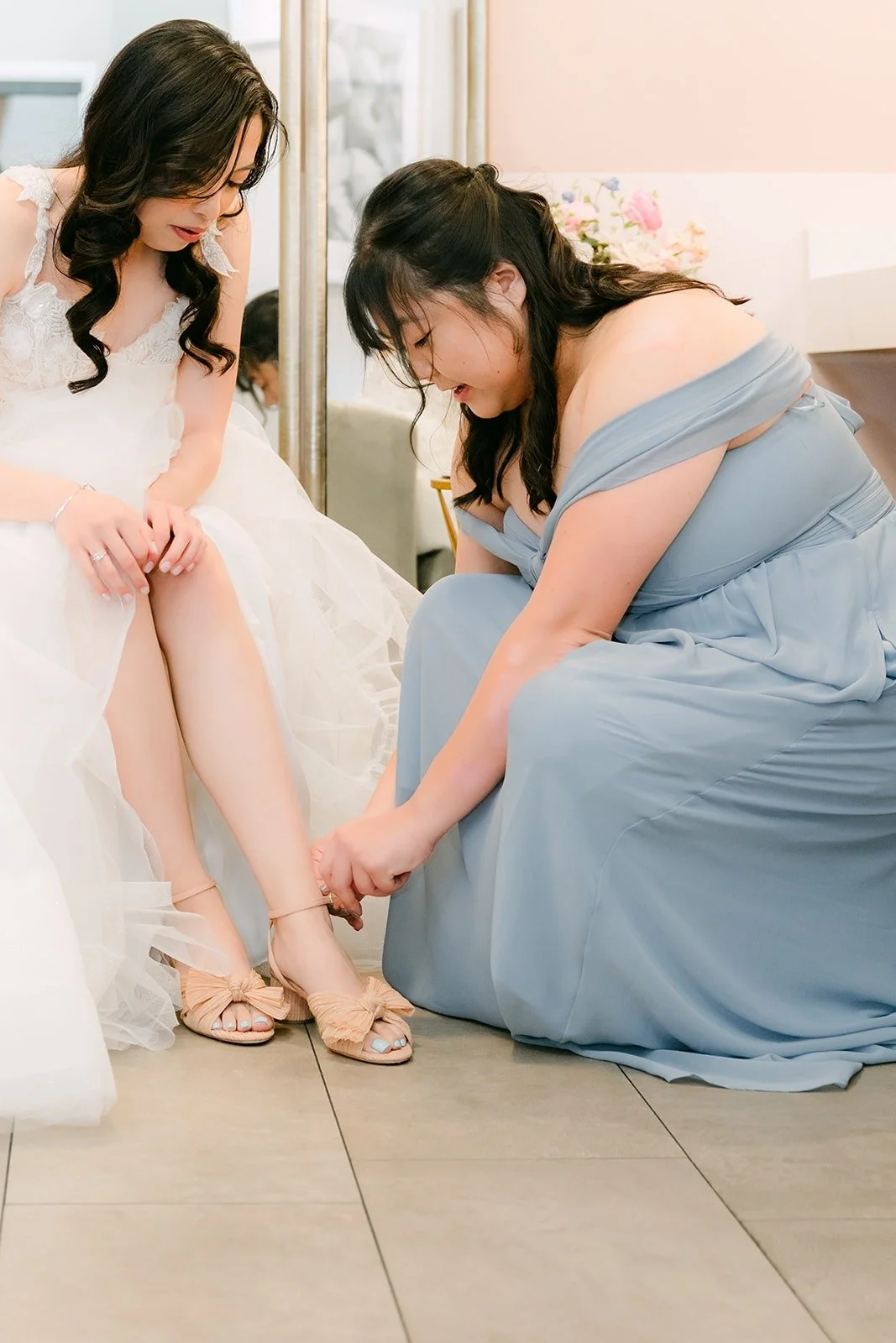 A bridesmaid in dusty blue helping Natalie into her wedding shoes in the bridal suite