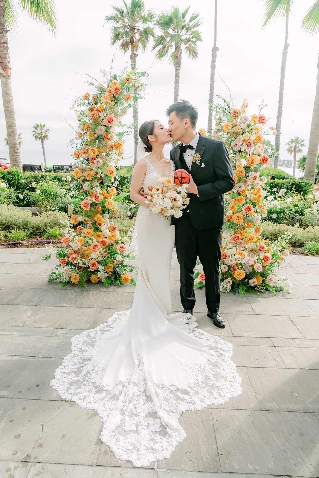 Angela and Eli kissing in front of the peach floral ceremony arch, her lace train spread out