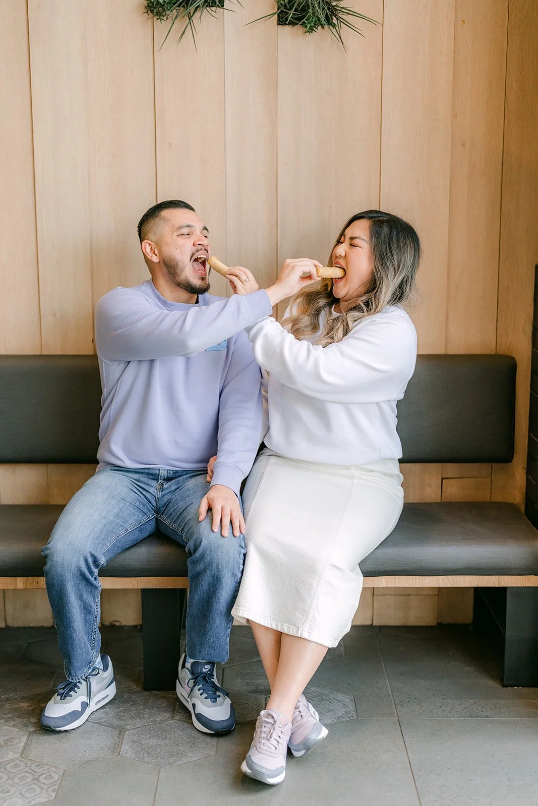 Ankush and Linh feed each other on a bench by the wood wall.