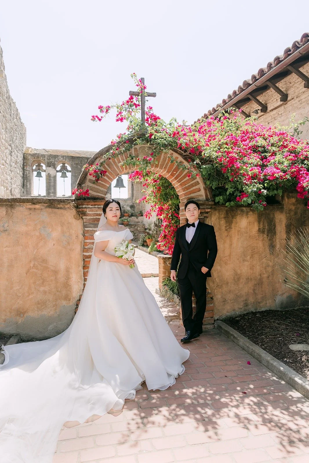 Chorok and Min Soo near a bougainvillea covered arch with a cross above it