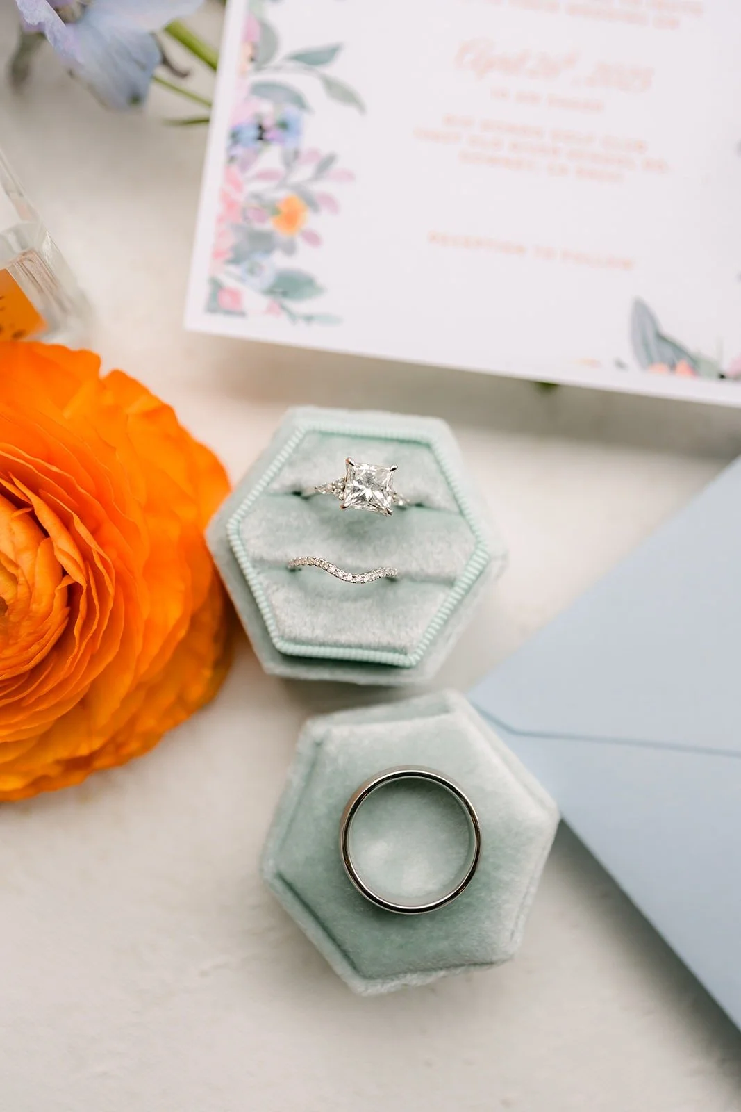 Two hexagon ring boxes in soft blue beside the invitation and orange ranunculus