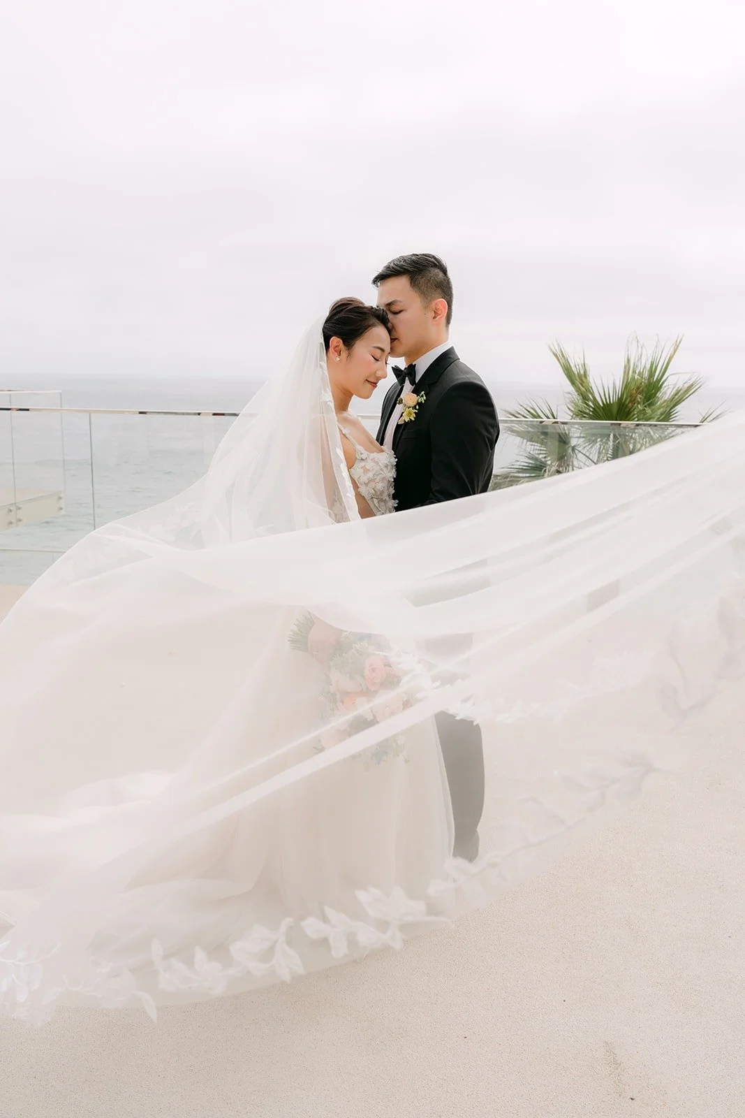 Kylie's veil blowing in the ocean breeze as Mario holds her close