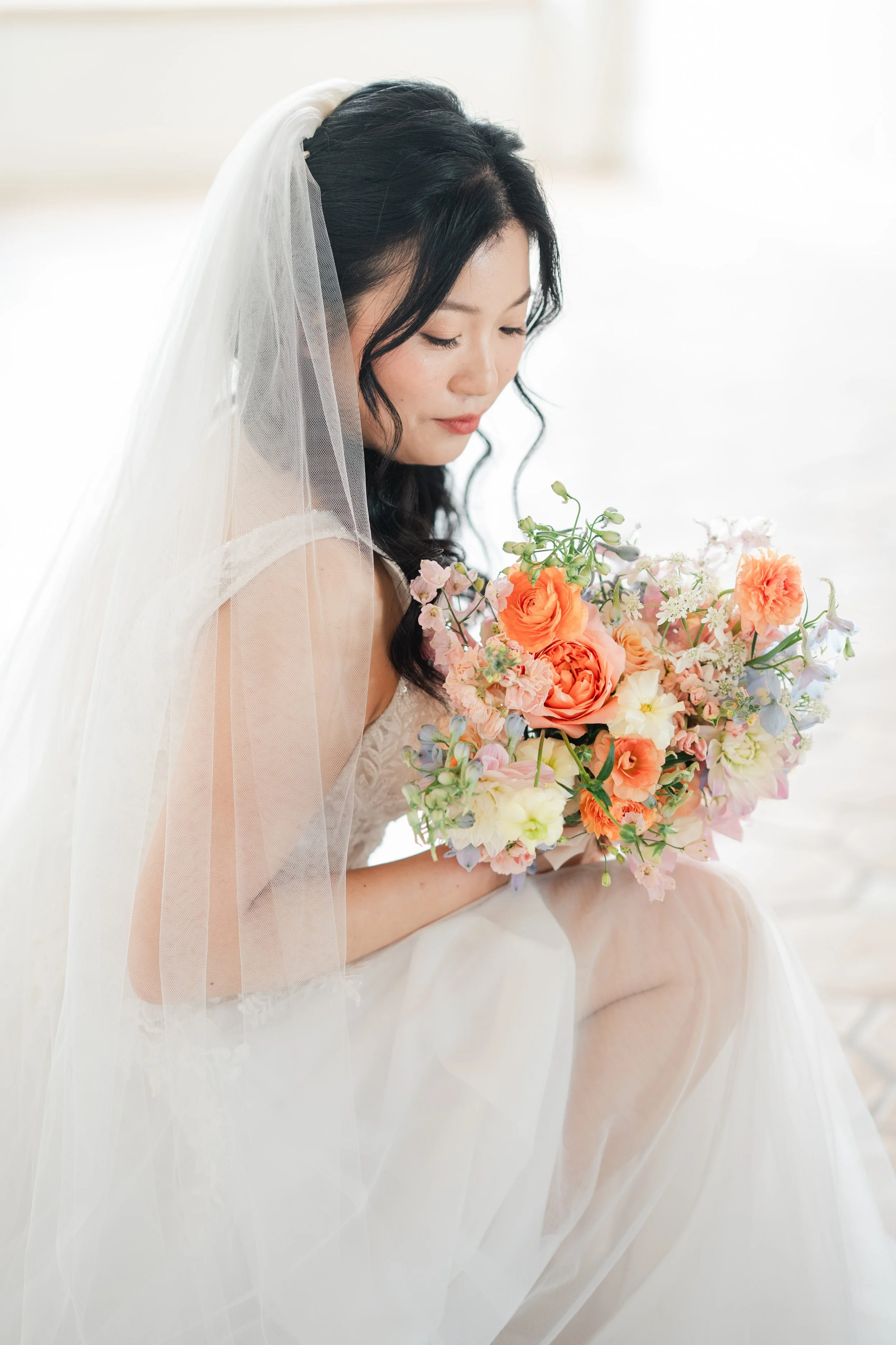 A bride with dark, wavy hair wearing a white wedding dress and veil, holding a bouquet of pink, orange, and white flowers, sitting with her eyes closed and a gentle expression.