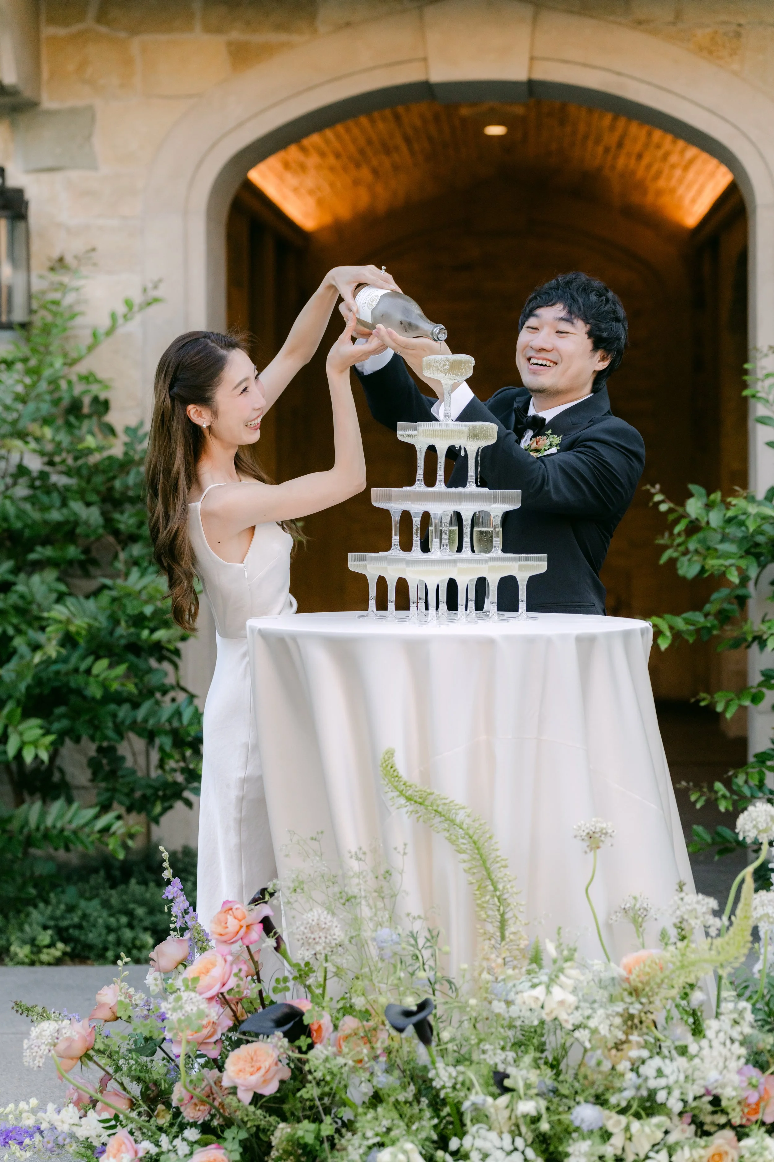 A bride and groom at a wedding celebration pouring champagne into a tower of glasses, surrounded by floral arrangements.