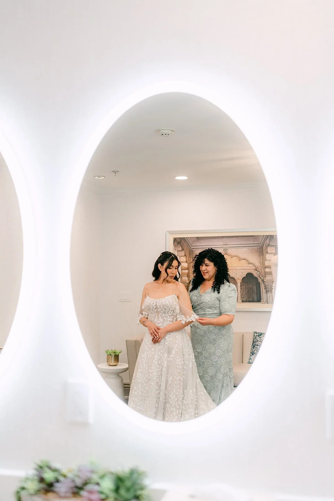 Fernanda and a bridesmaid reflected in a round lit mirror.