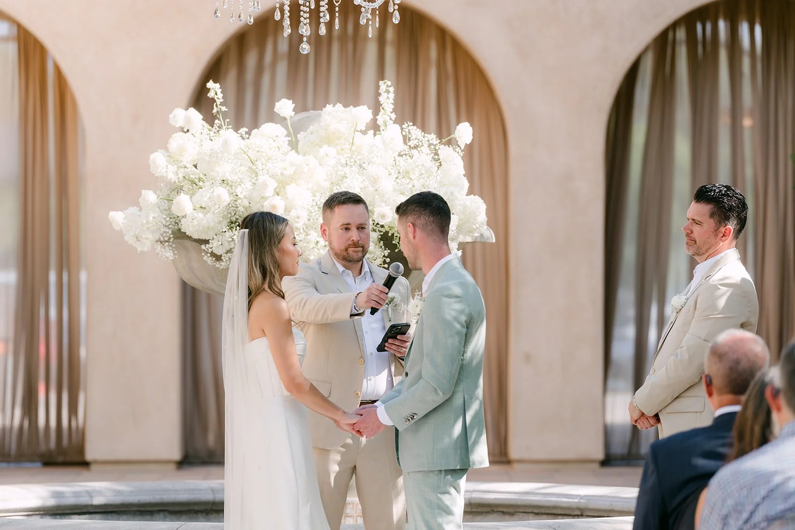 Kieu and Anthony beneath a draped arch with tall white florals as Nick officiates