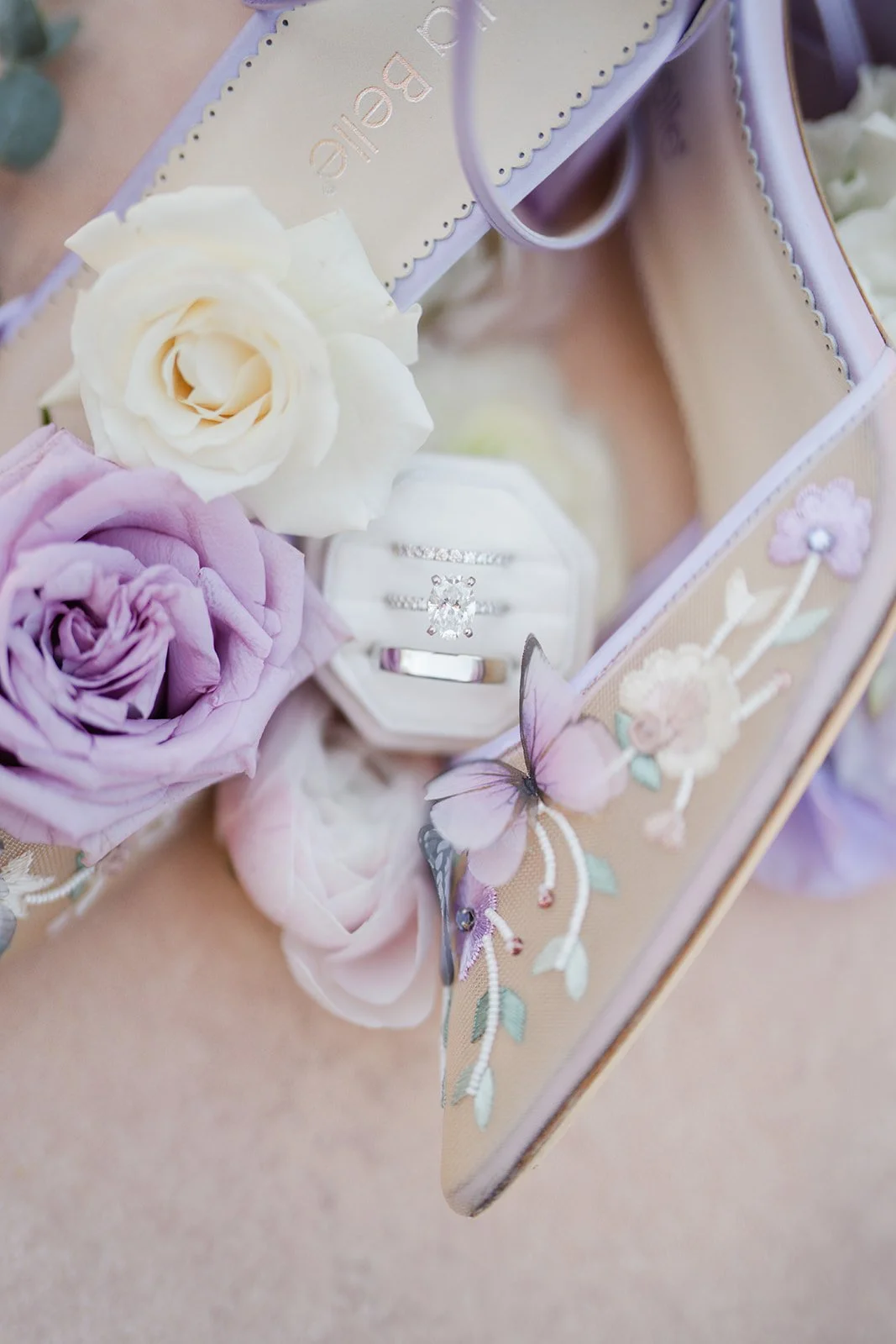 Bridal shoes with butterfly details beside lavender roses and the rings