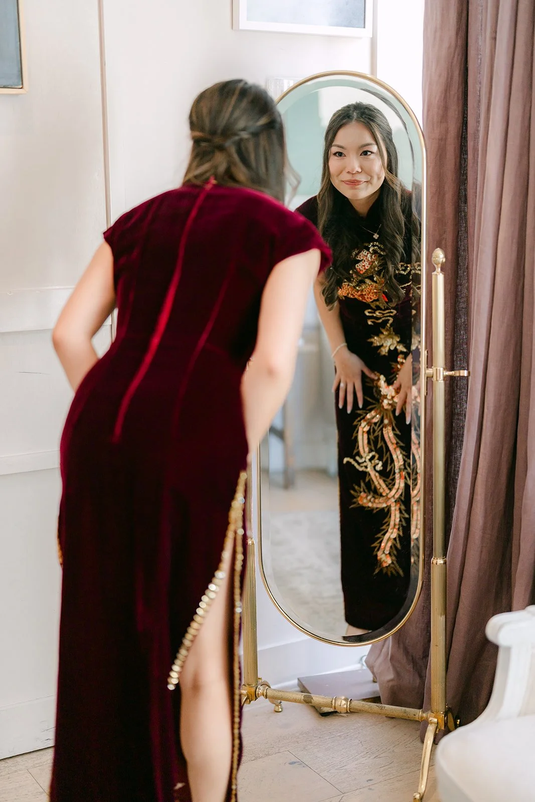 A family member helping Jadee with her qipao by the arched mirror