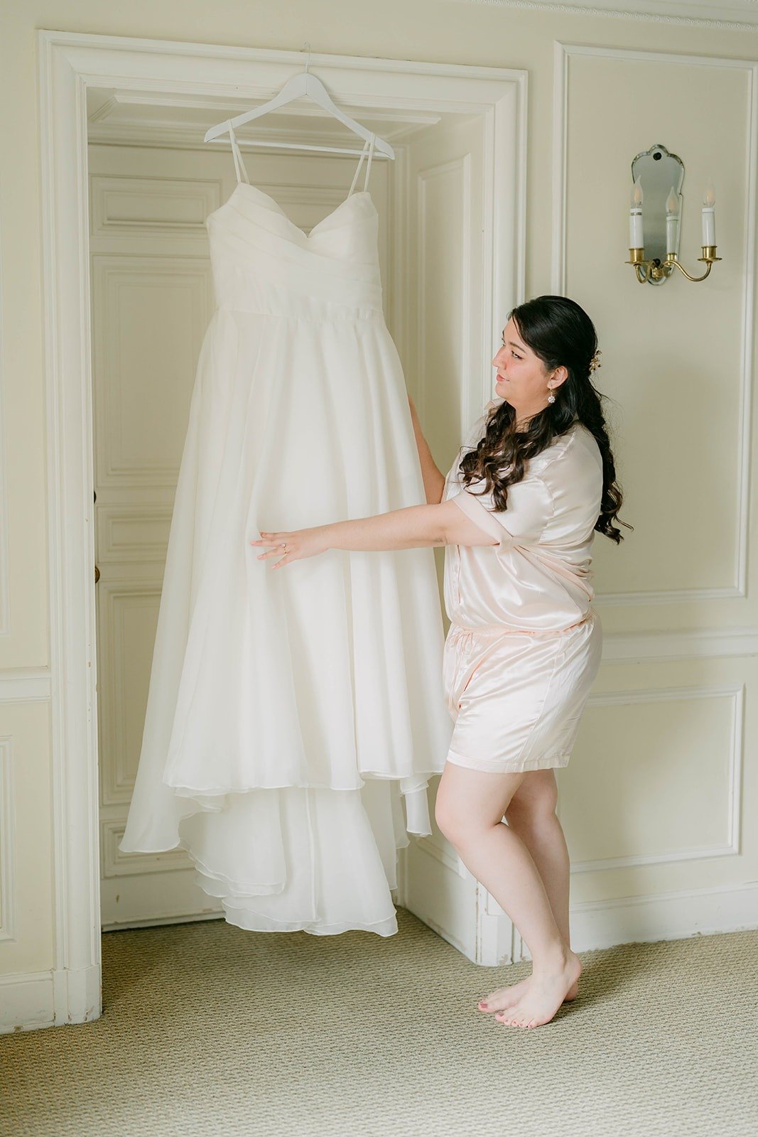 Saira in her pajamas beside her wedding dress hanging in the Mezzanine at The Athenaeum