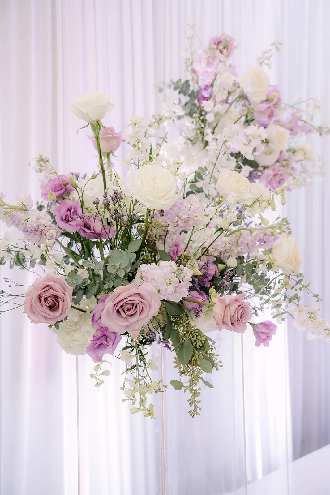 A tall arrangement of lavender and white blooms against white draping