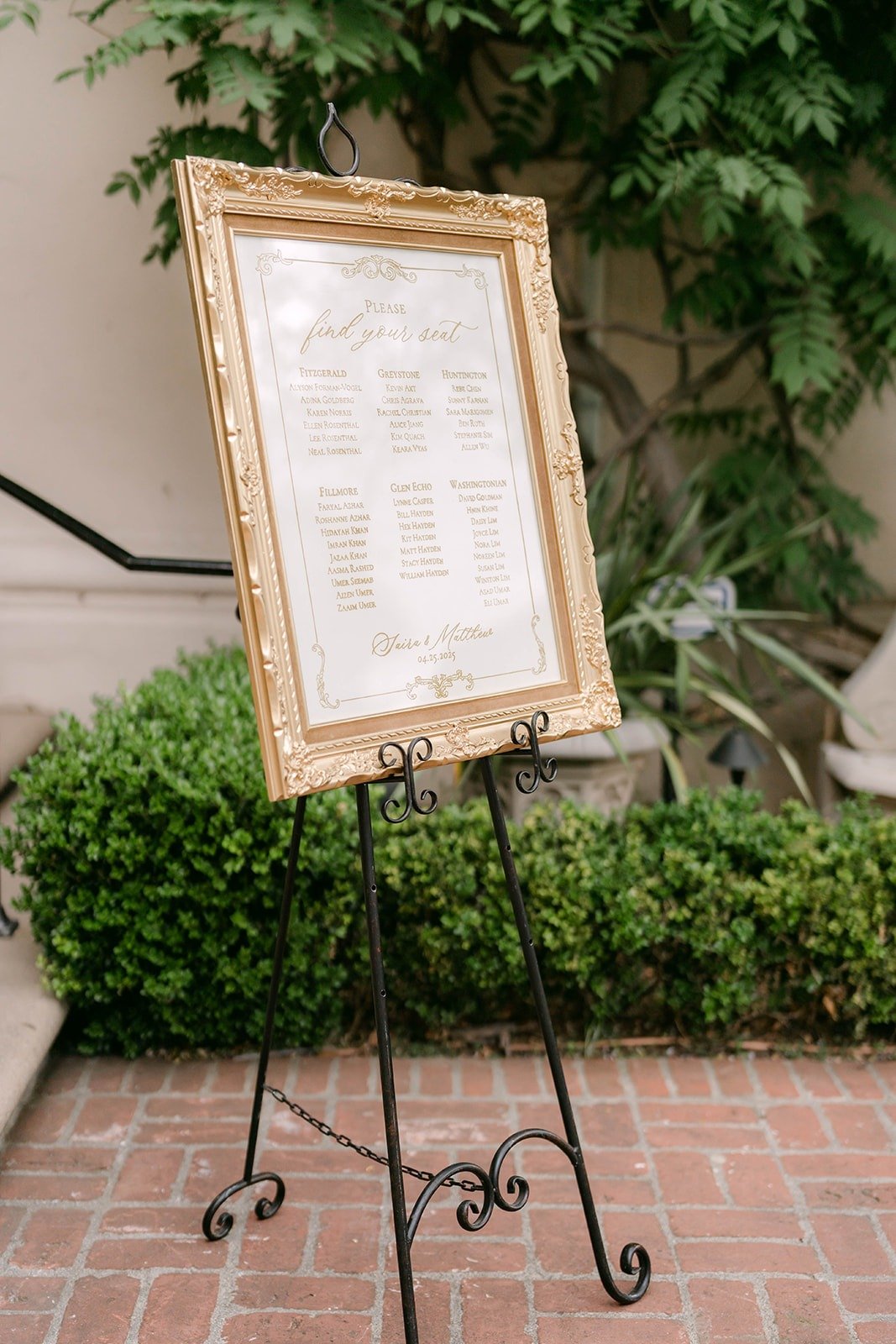 The seating chart in a gold frame on an easel beside green hedges