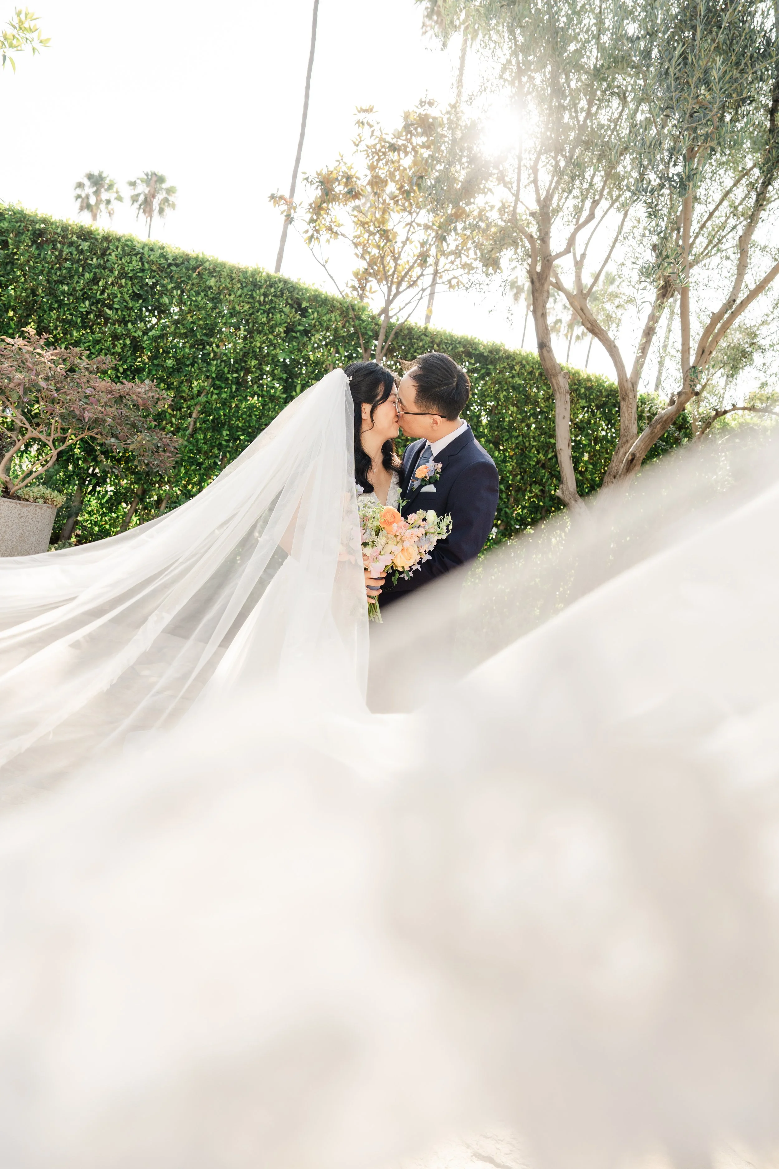 A bride and groom share a kiss outdoors, with trees, a green hedge, and bright sunlight in the background, as the bride's long veil flows around them.