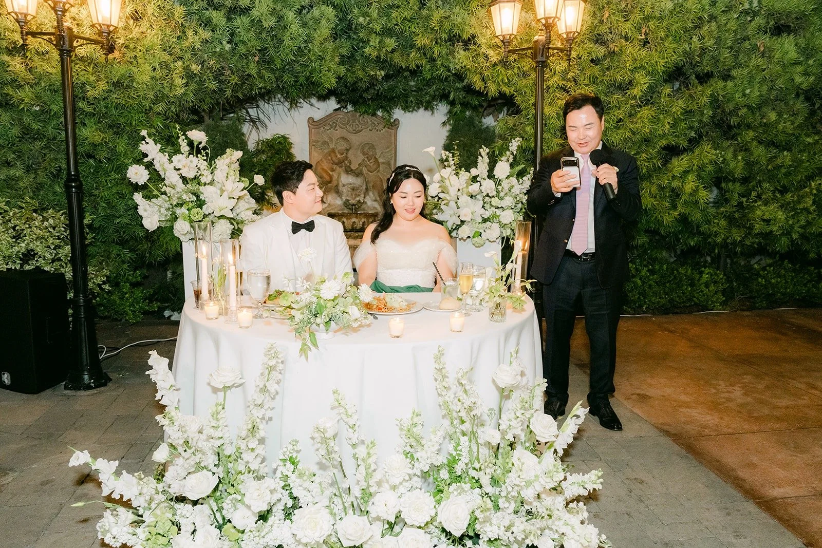 A guest speaking beside the sweetheart table as Chorok and Min Soo listen