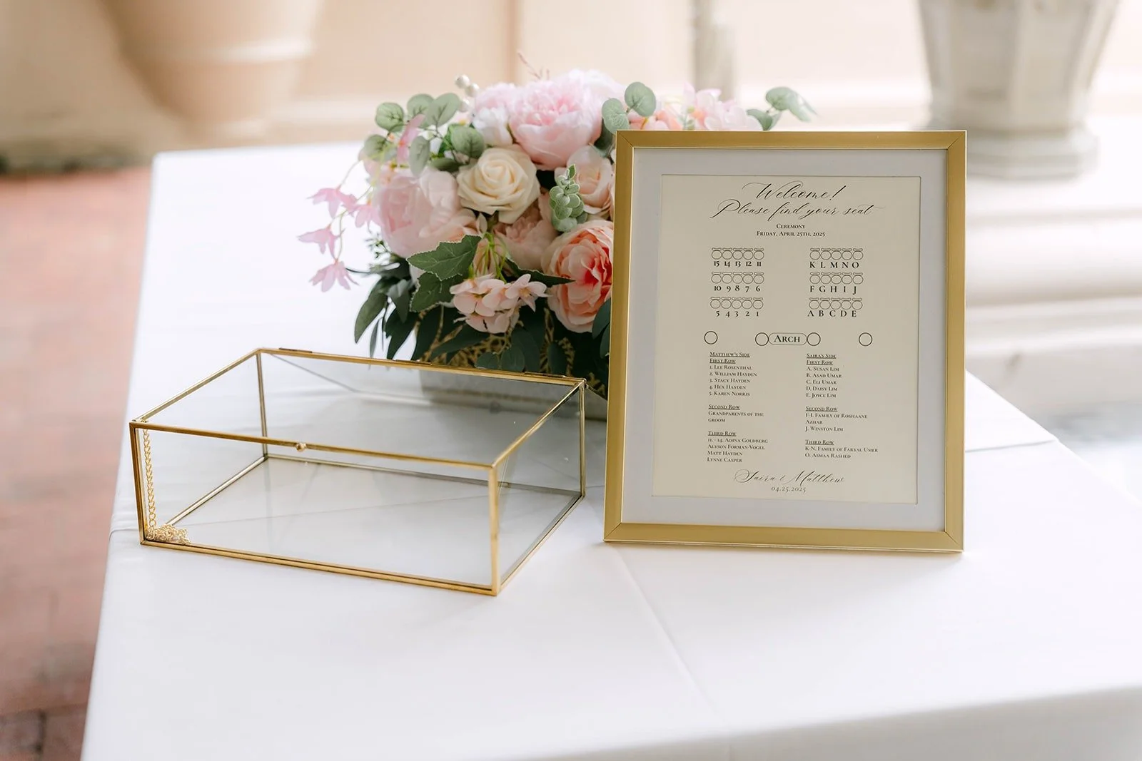 A gold frame program and a gold card box beside blush flowers at the ceremony entrance