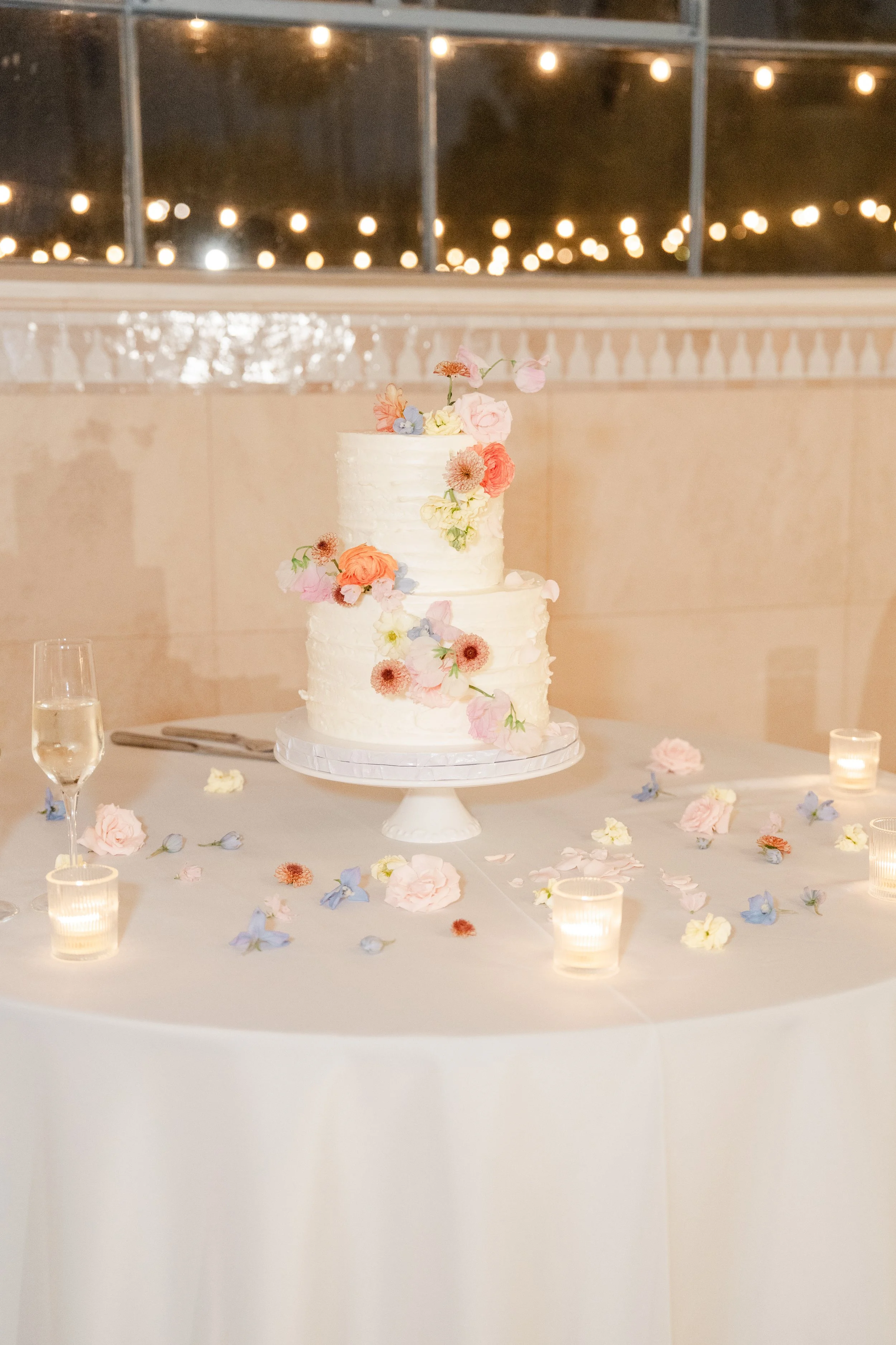 A white, two-tiered wedding cake decorated with pink, peach, and light purple flowers on a table, surrounded by scattered flower petals and small candles.