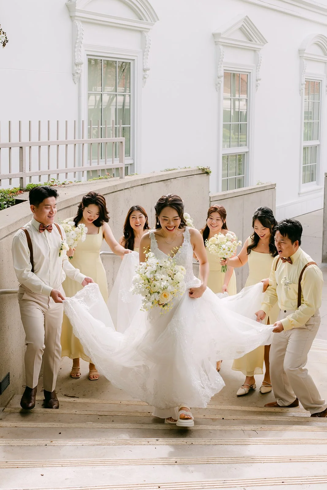 Bridesmaids fluffing Jenny's train on the steps by a white building.
