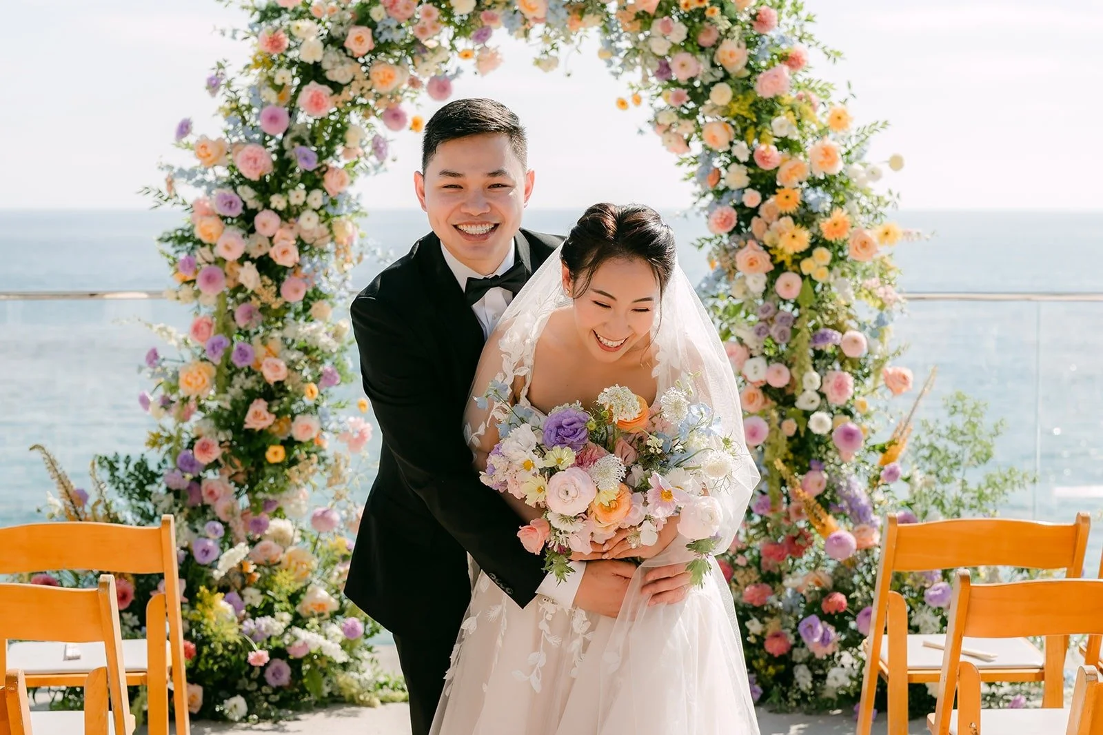Mario hugging Kylie from behind under the pastel floral arch, photographed by One and Only Production