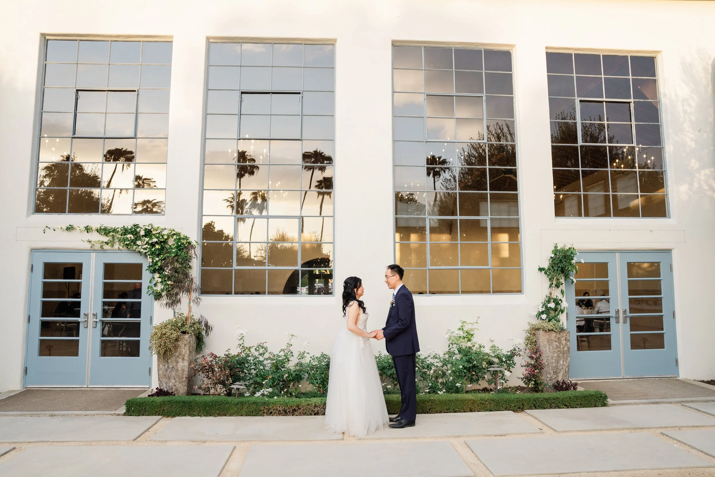 A bride and groom holding hands and facing each other outside in front of a modern building with large windows reflecting the sky and palm trees.