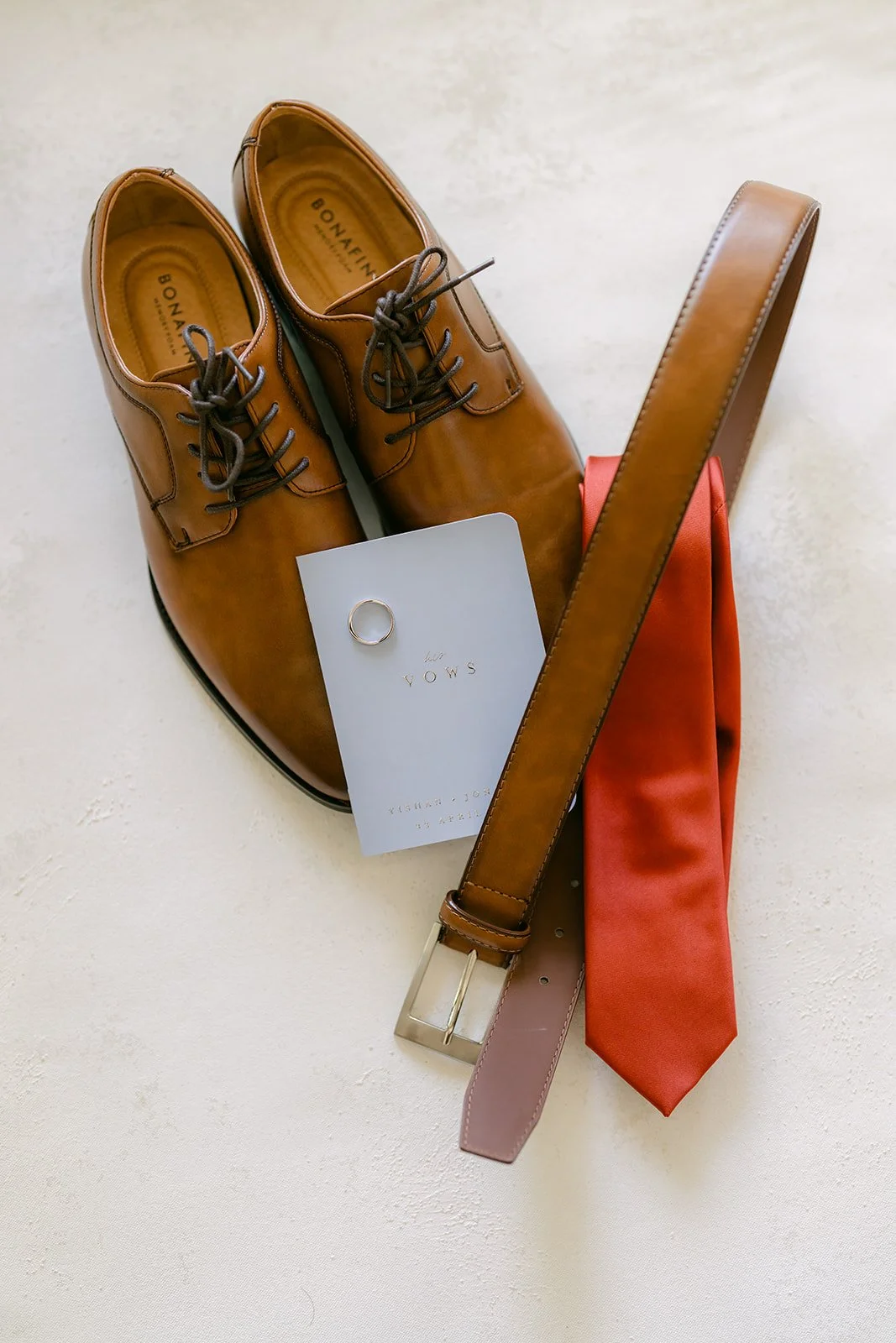 Groom's brown leather shoes, belt and rust tie laid out on white