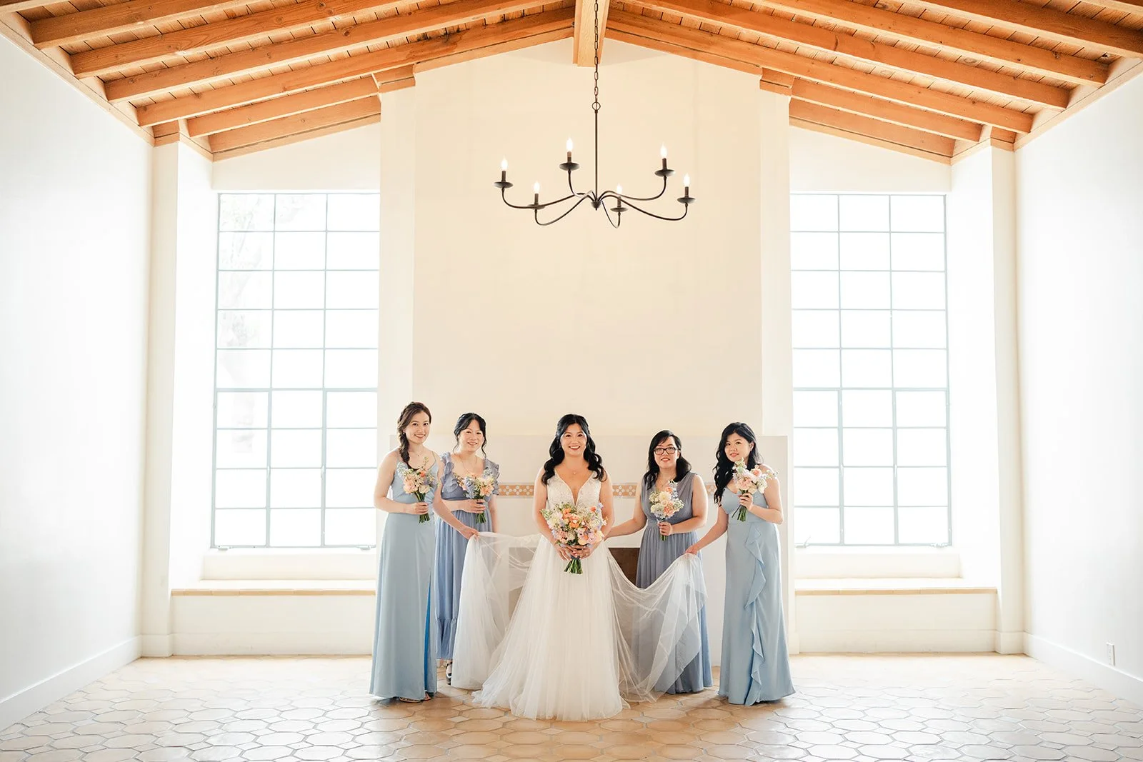 Bride and bridesmaids in dusty blue in a sunlit hall with arched windows and wood beams