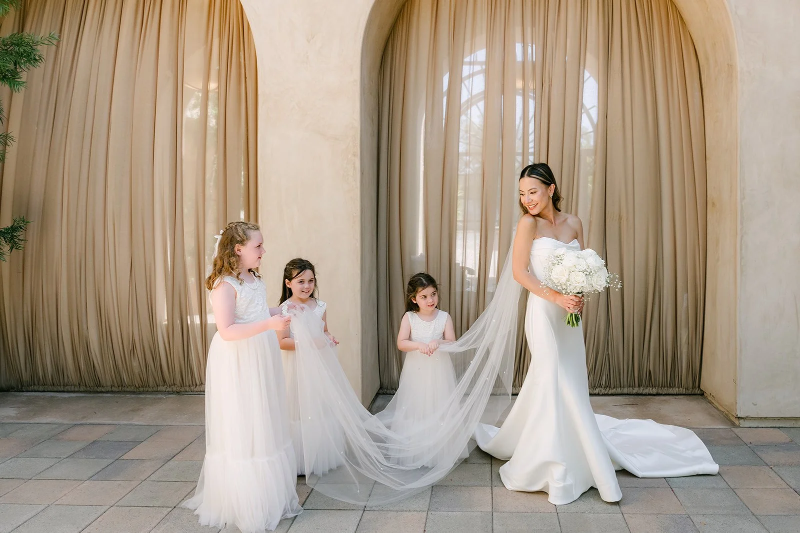 Bride holding her veil with the flower girls beside the arched curtains
