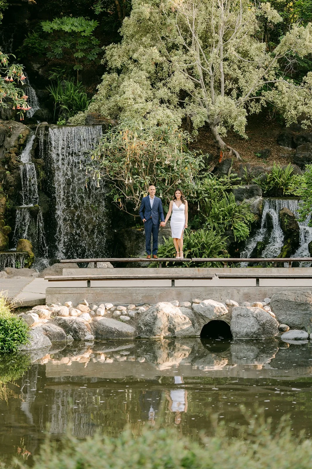 Francesca and Daniel stand on a stone path in front of the waterfall at the LA Arboretum.