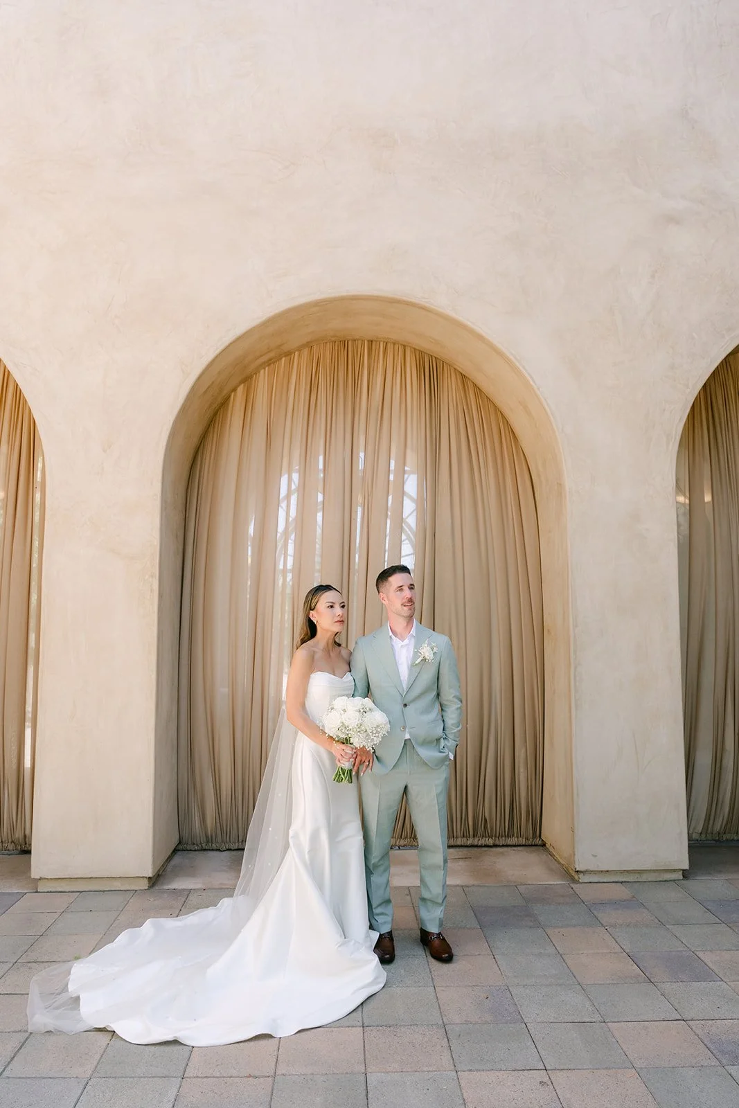 Wide portrait of the couple in front of a curtained arch with her train spread out
