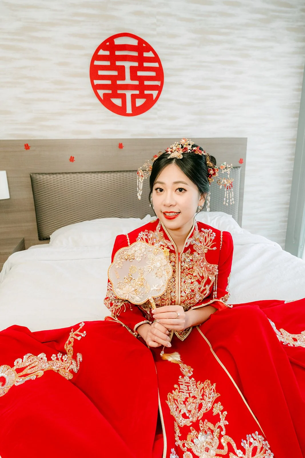 A bride waiting on the bed in her qun kwa holding a fan before the groom arrives.