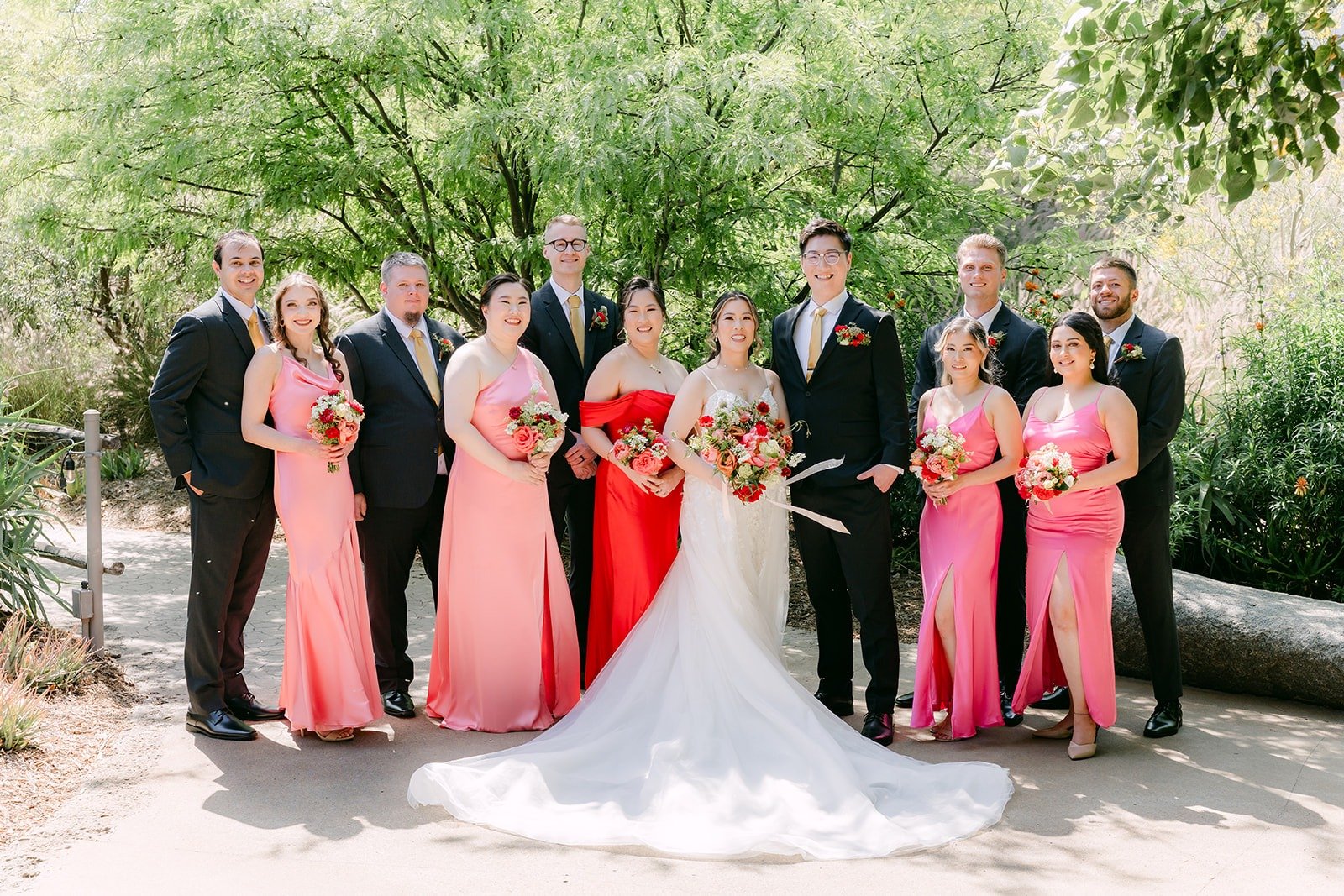 The full wedding party in pink, red and black suits around Joanne and Zach in the garden