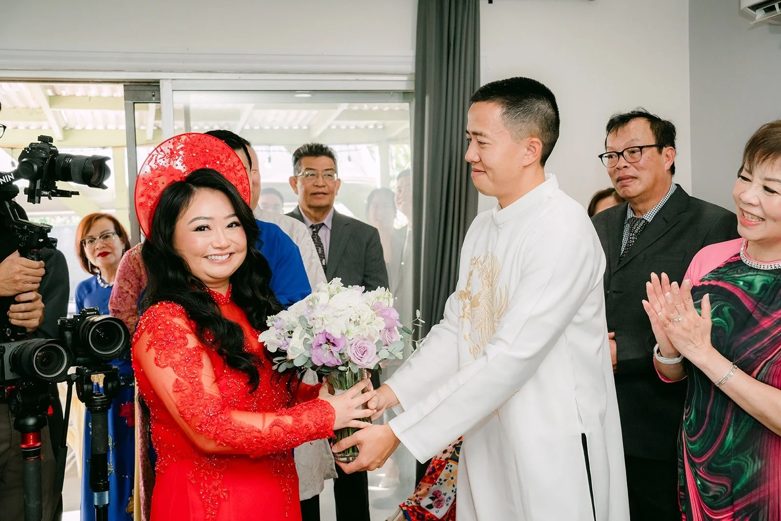 Kevin, in a white ao dai, hands Jennifer her bouquet in her red ao dai and headpiece