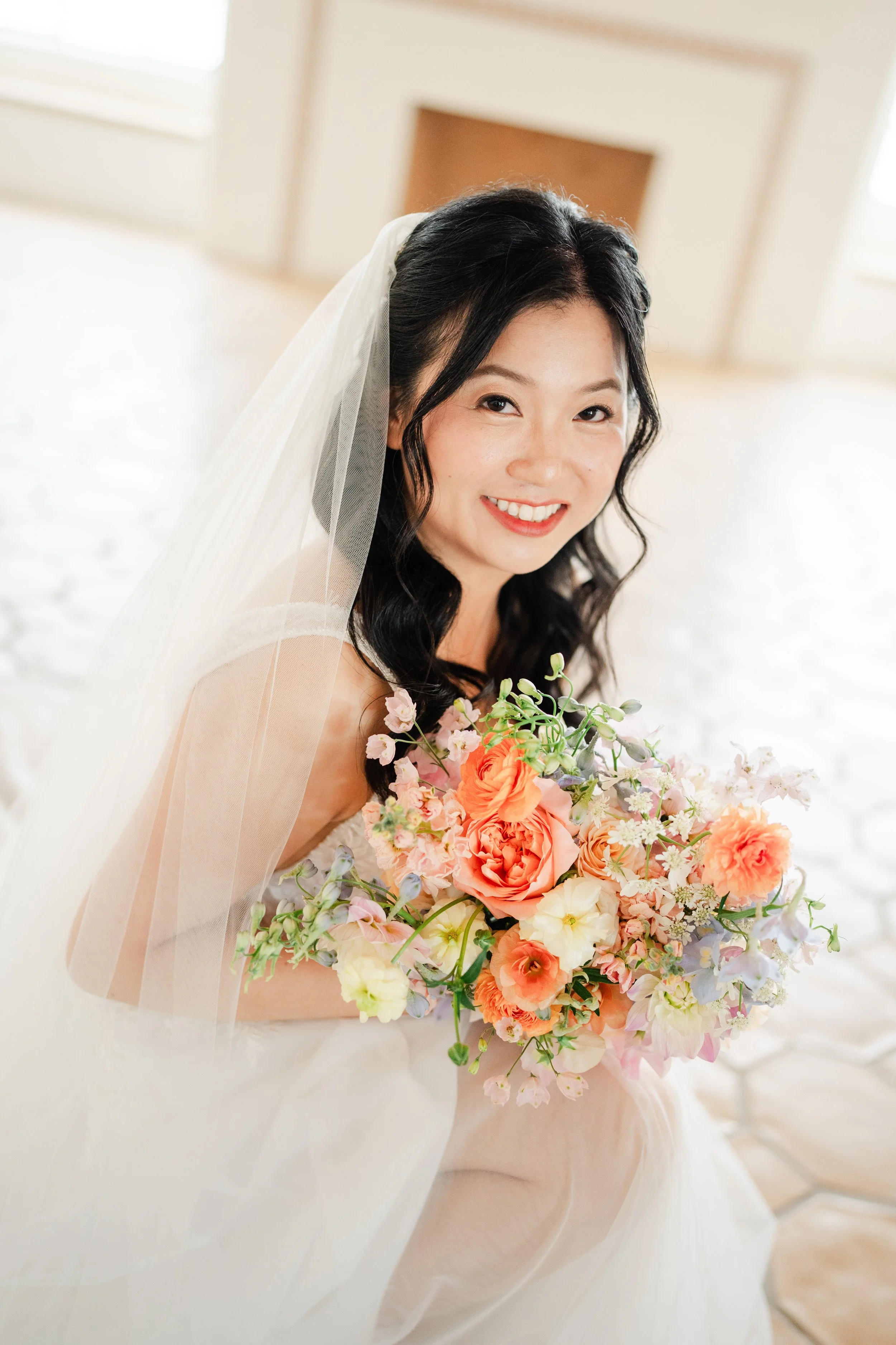 A smiling bride with dark hair and a veil, holding a colorful bouquet of pink, orange, and white flowers, indoors near a fireplace.