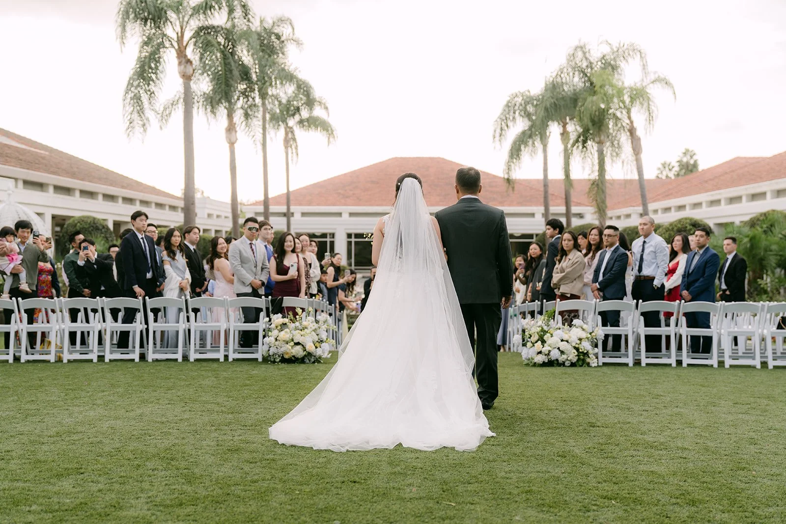 Jenny walking down the aisle with her dad, framed by palm trees.