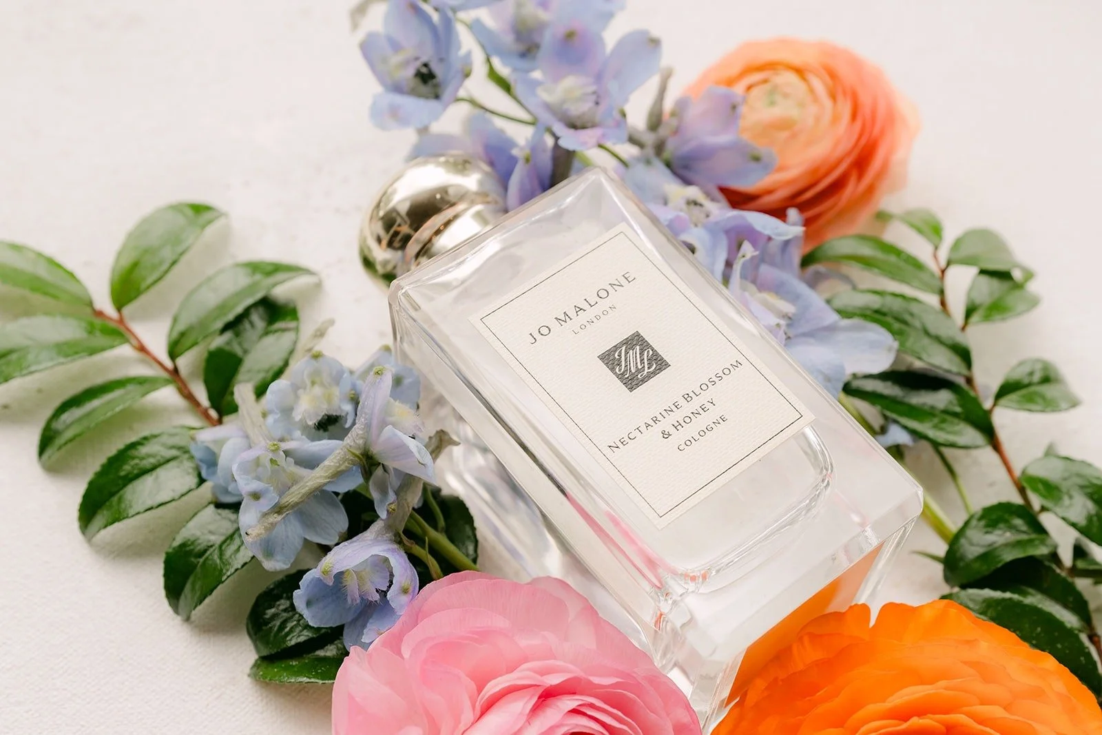 A perfume bottle styled among blue hydrangea, orange ranunculus and greenery