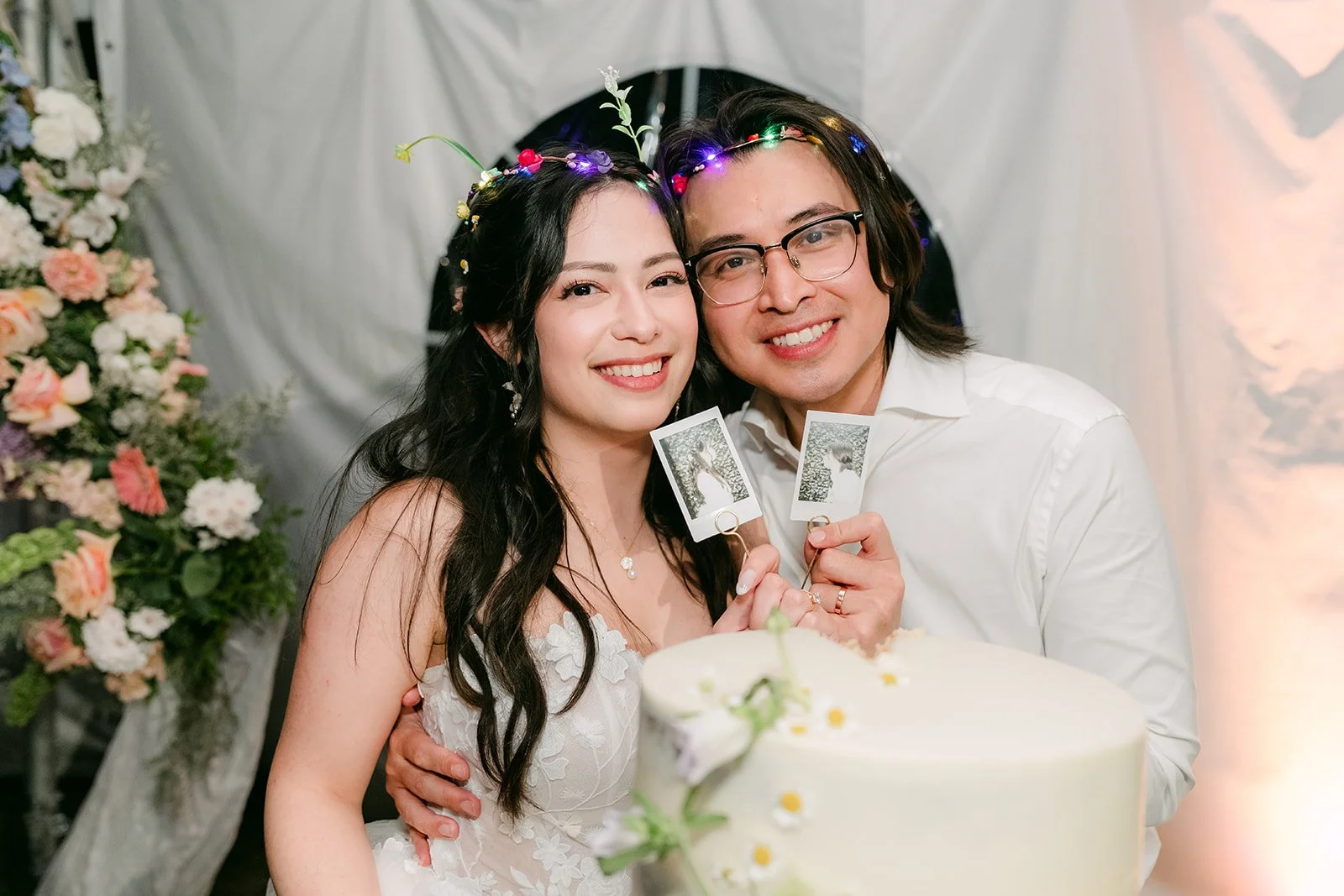 Newlyweds in light-up flower crowns smiling beside their cake