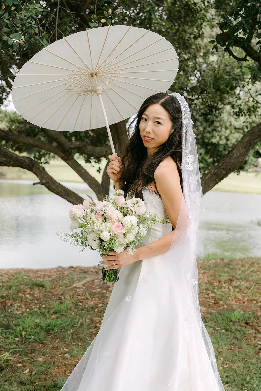 Kristine holding a white paper parasol and her bouquet by the water