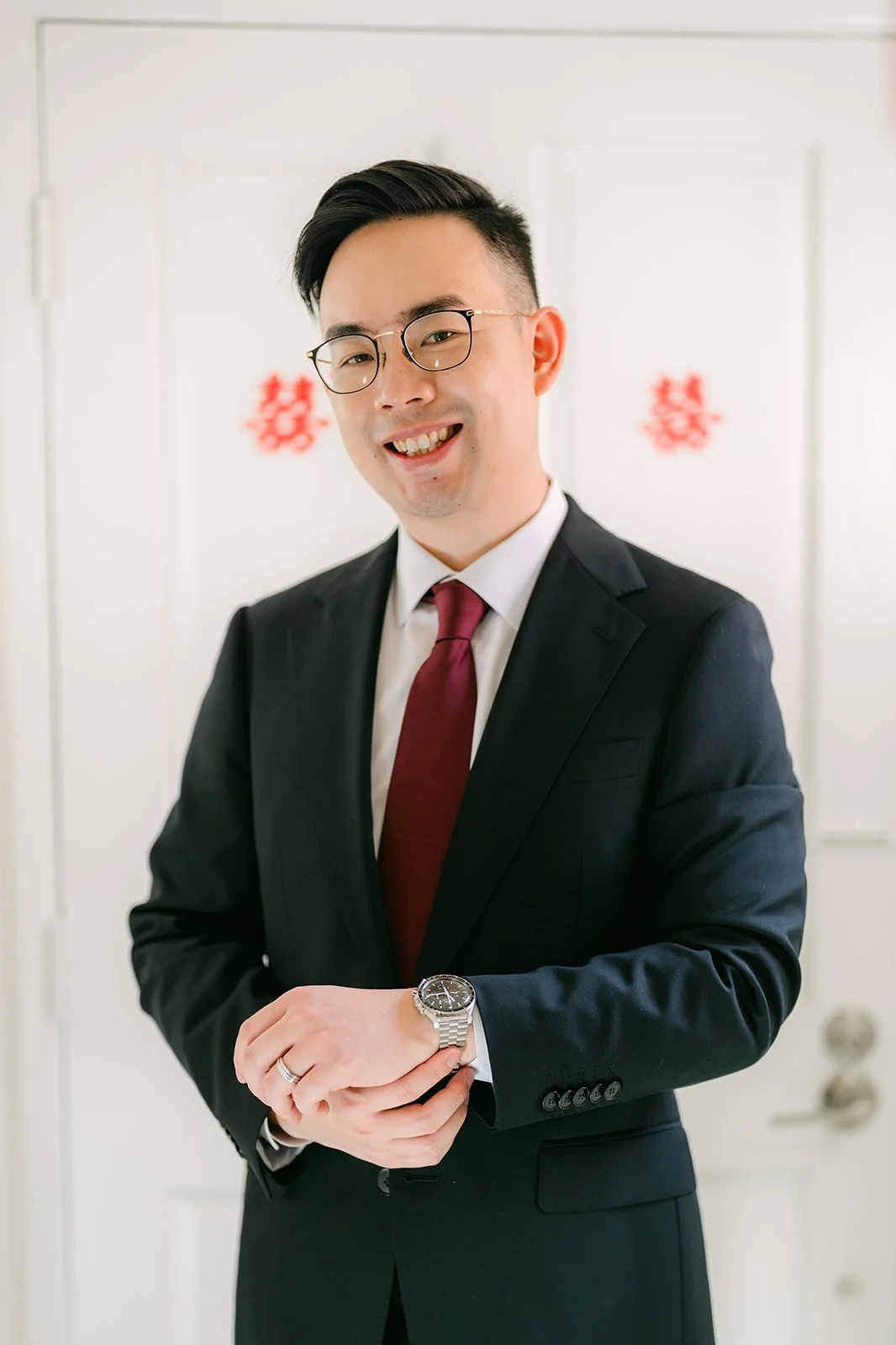 A groom in a dark suit and red tie standing in the hallway before the door game.