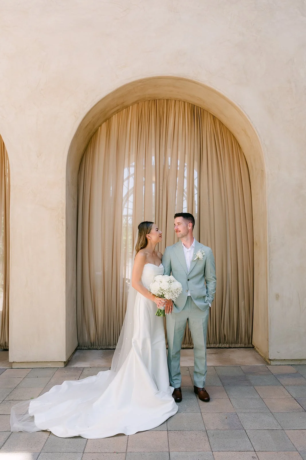 Couple standing beneath a curtained arch with her train spread on the pavers