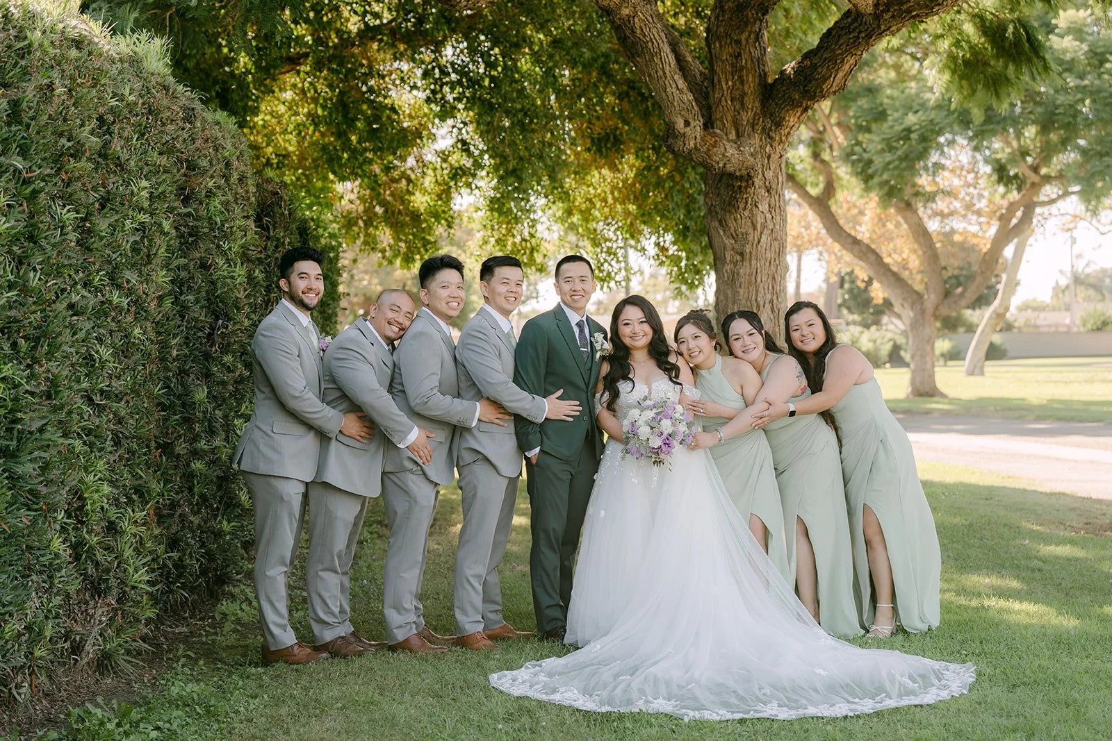 The full wedding party lines up by the hedge at Garden Grove Park
