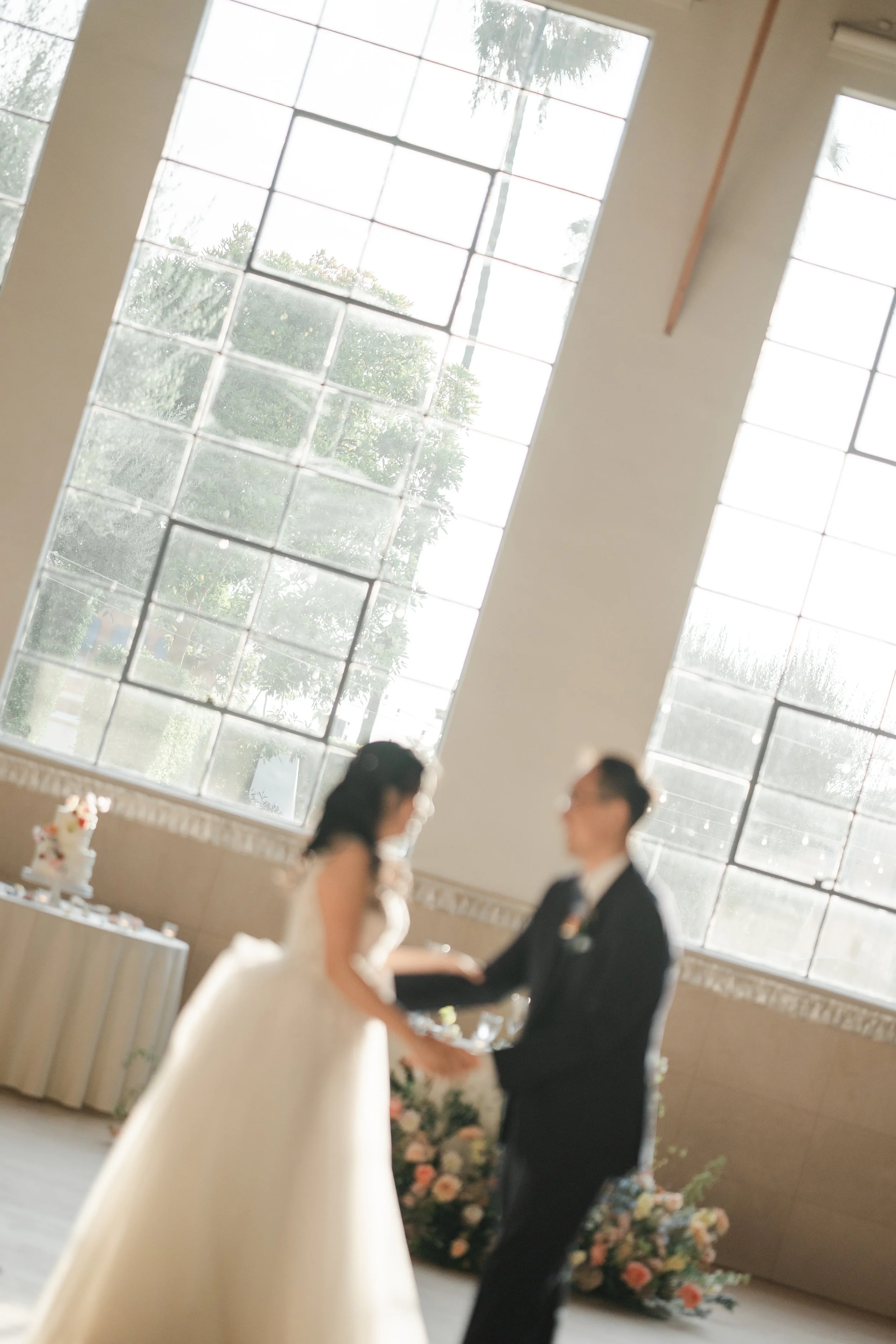 A bride and groom holding hands and facing each other during a wedding ceremony, standing inside a room with large windows and a view of trees outside, with floral arrangements in the background.
