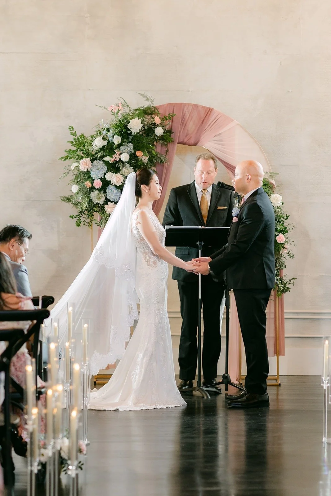 Elisa and Brandon hold hands in front of the floral arch as the ceremony begins