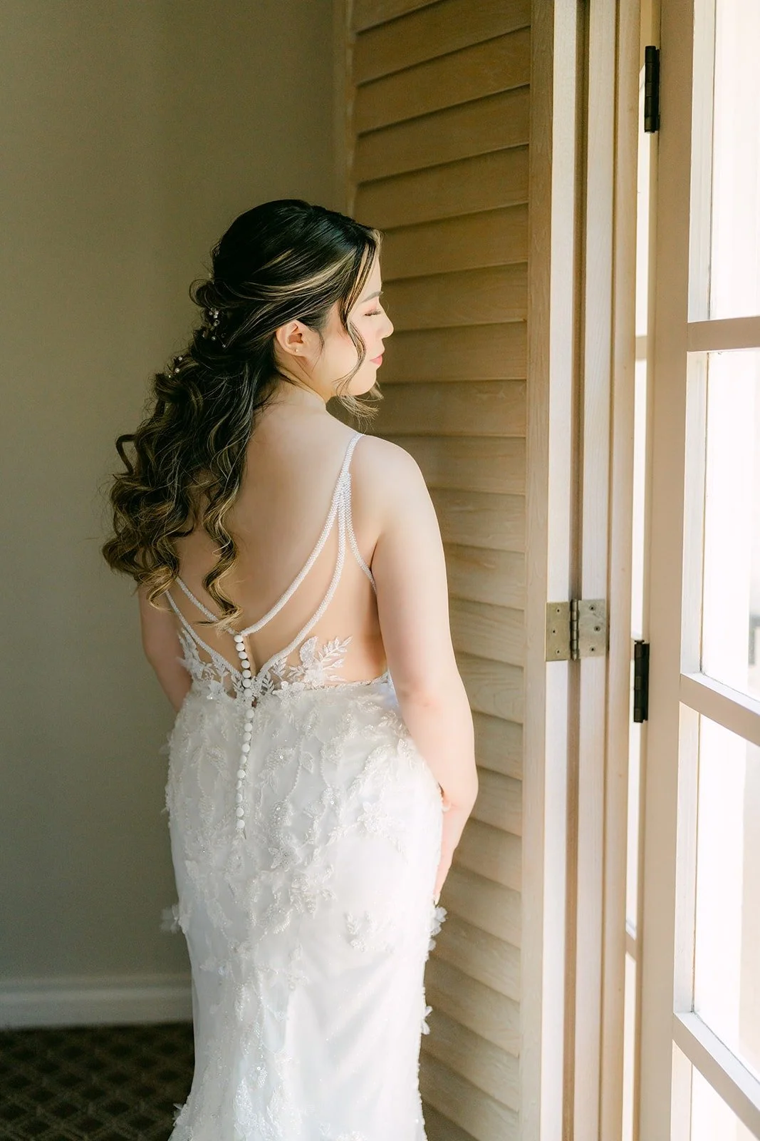 Joanne in her gown with its open back and crossed straps beside the window