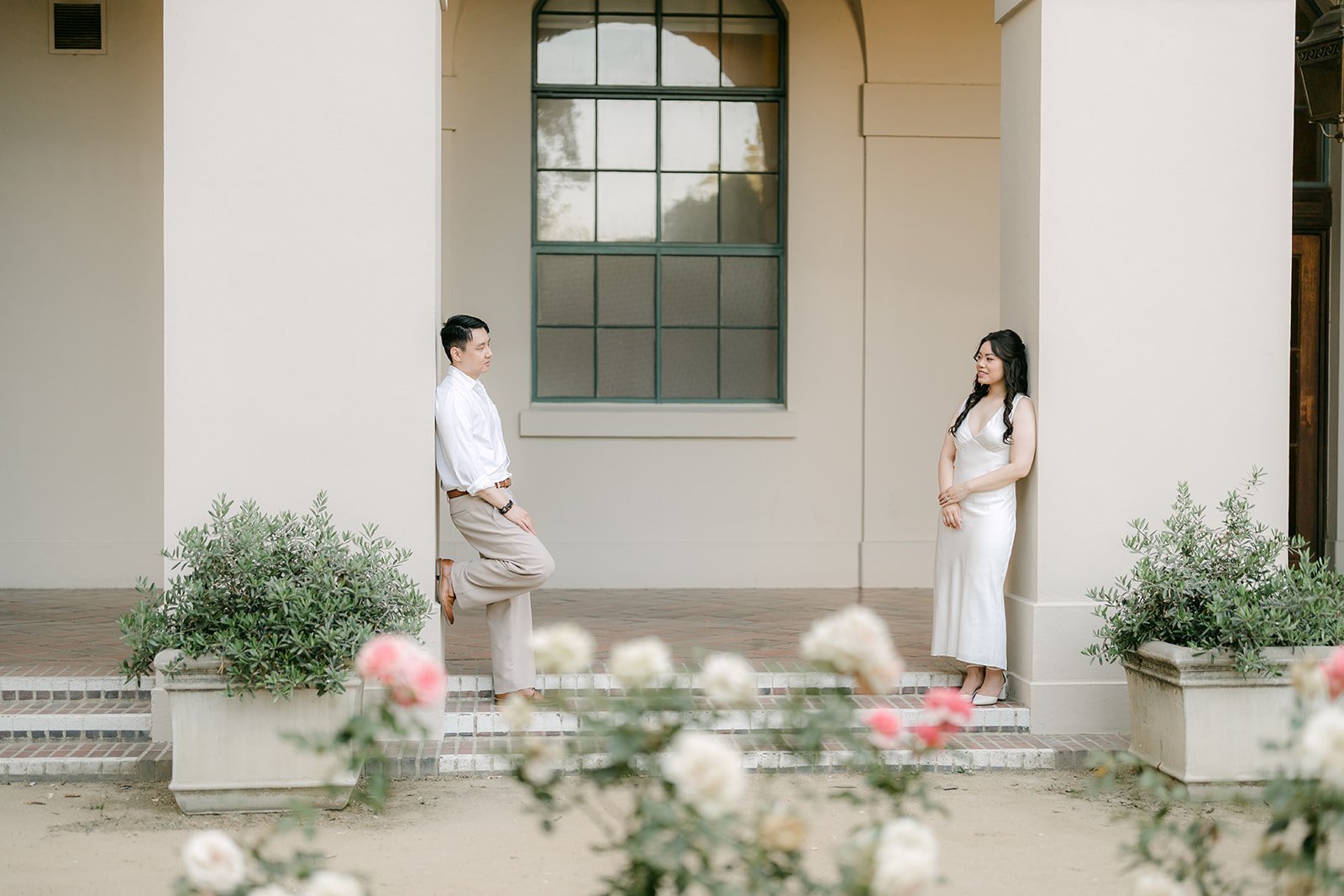 Emily and James stand below an arched window lined with potted plants.