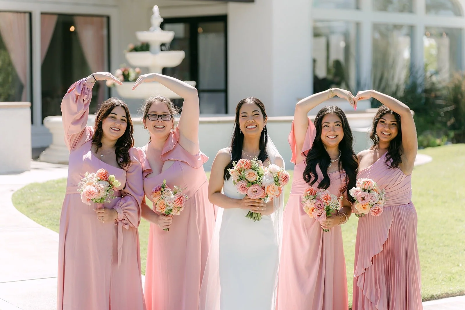 Stephanie's bridesmaids making heart hands around her in front of the fountain