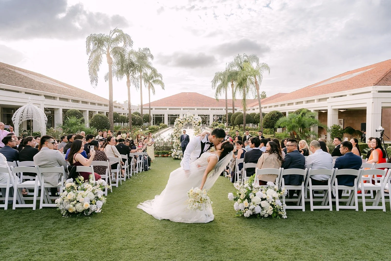 Wide view of Jenny and Ryan's dip kiss on the lawn with palms behind them, photographed by One and Only Production.