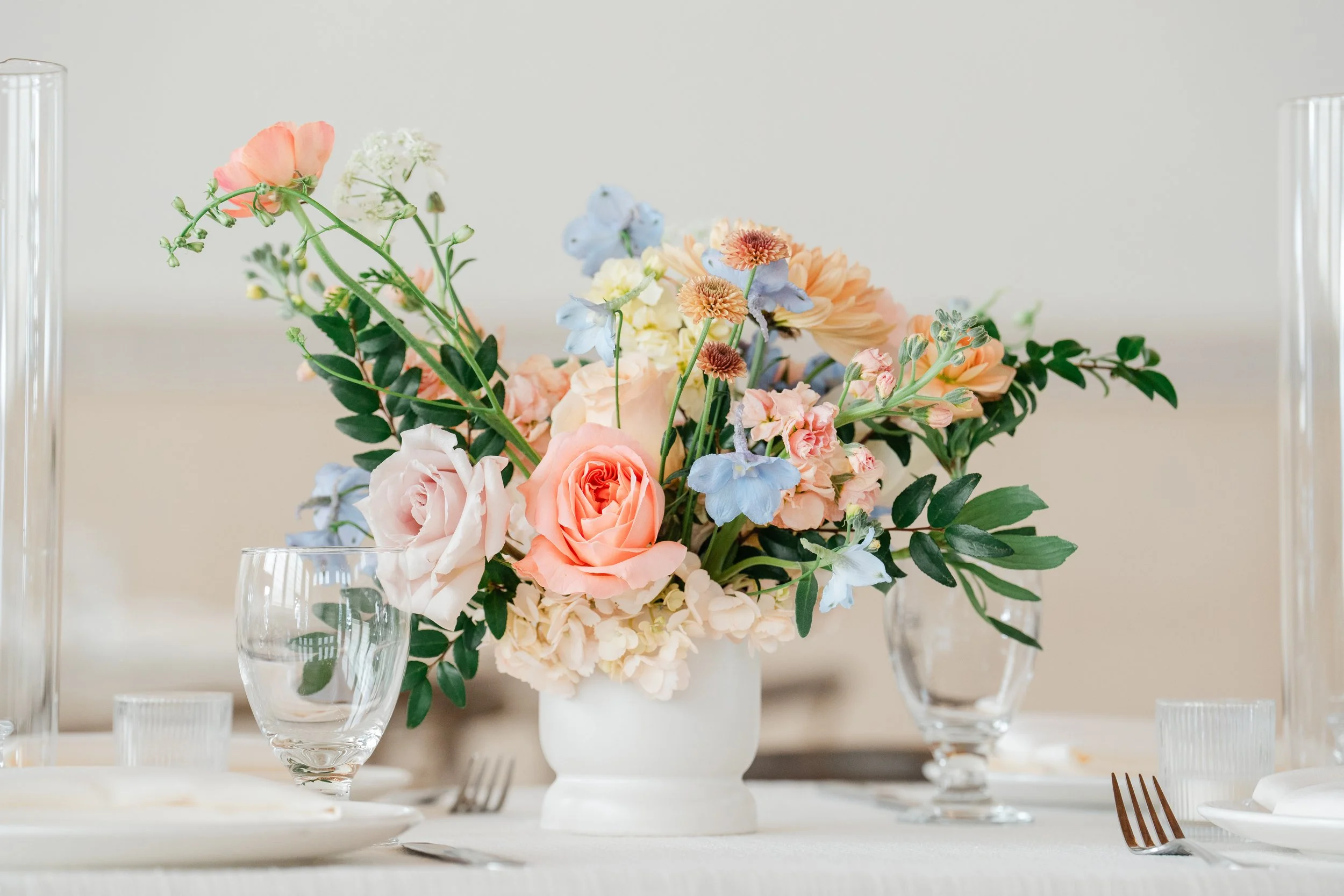 A floral centerpiece with pink, peach, and blue flowers on a white table set with glassware and dinnerware, with a neutral background.