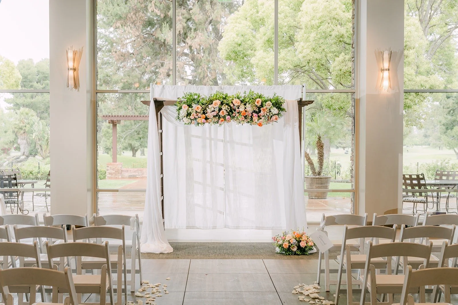 The white draped ceremony arch topped with peach and pink flowers in front of tall windows